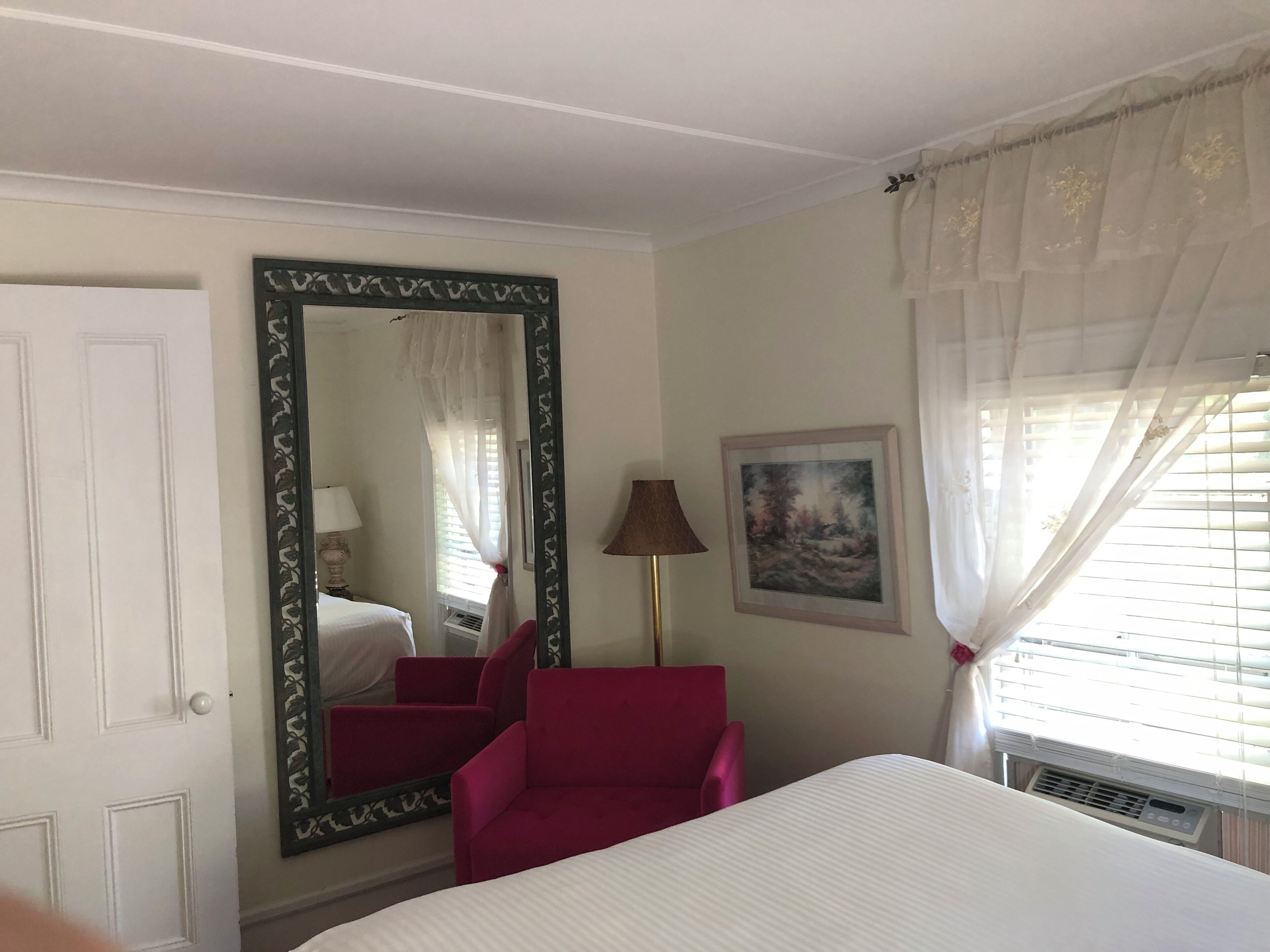 Una camera d'albergo con un letto, una poltrona rossa, un grande specchio, un quadro e una finestra con tende.