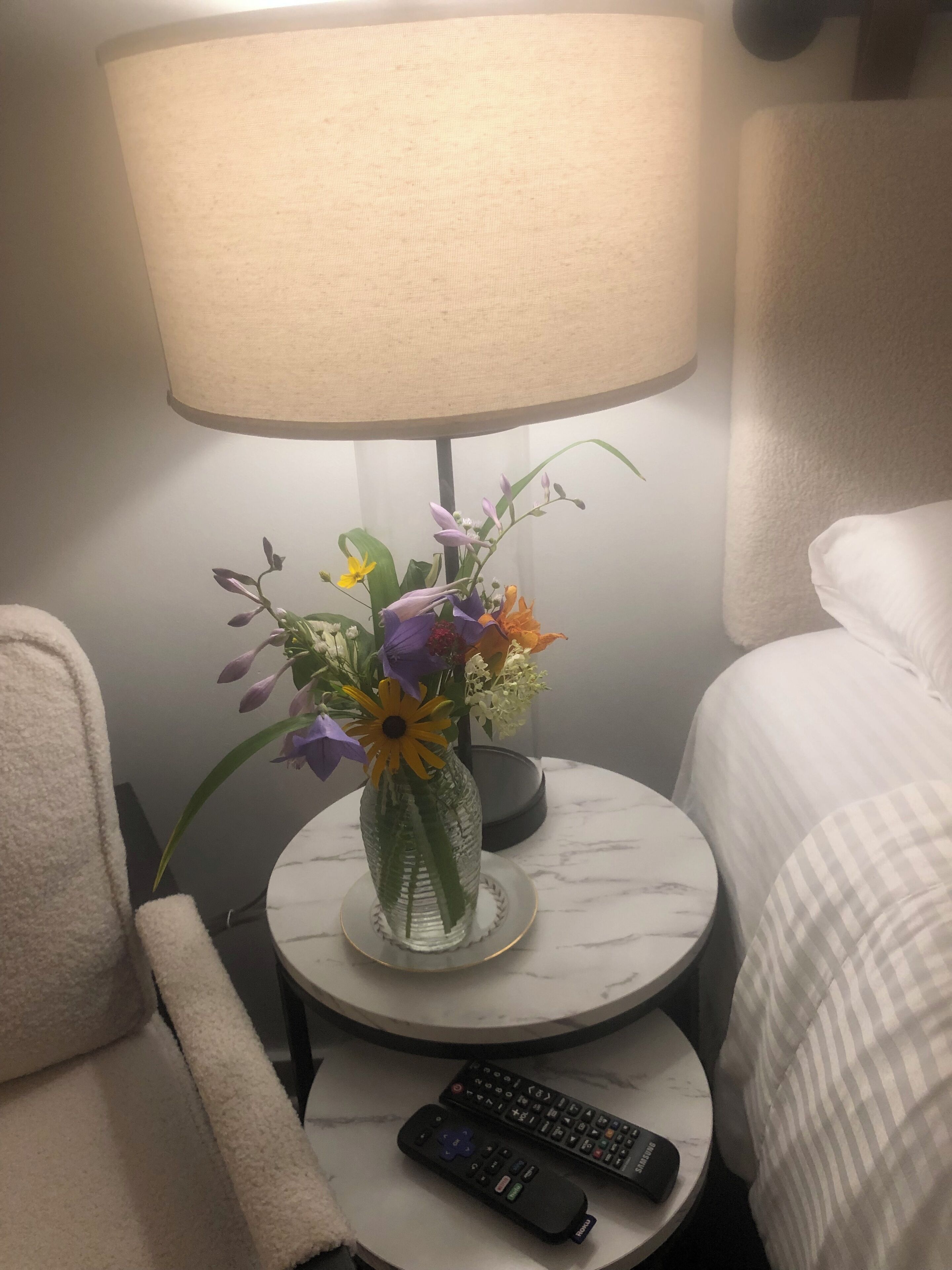 A hotel room with a round table, a lamp, a vase with flowers, and a remote control.
