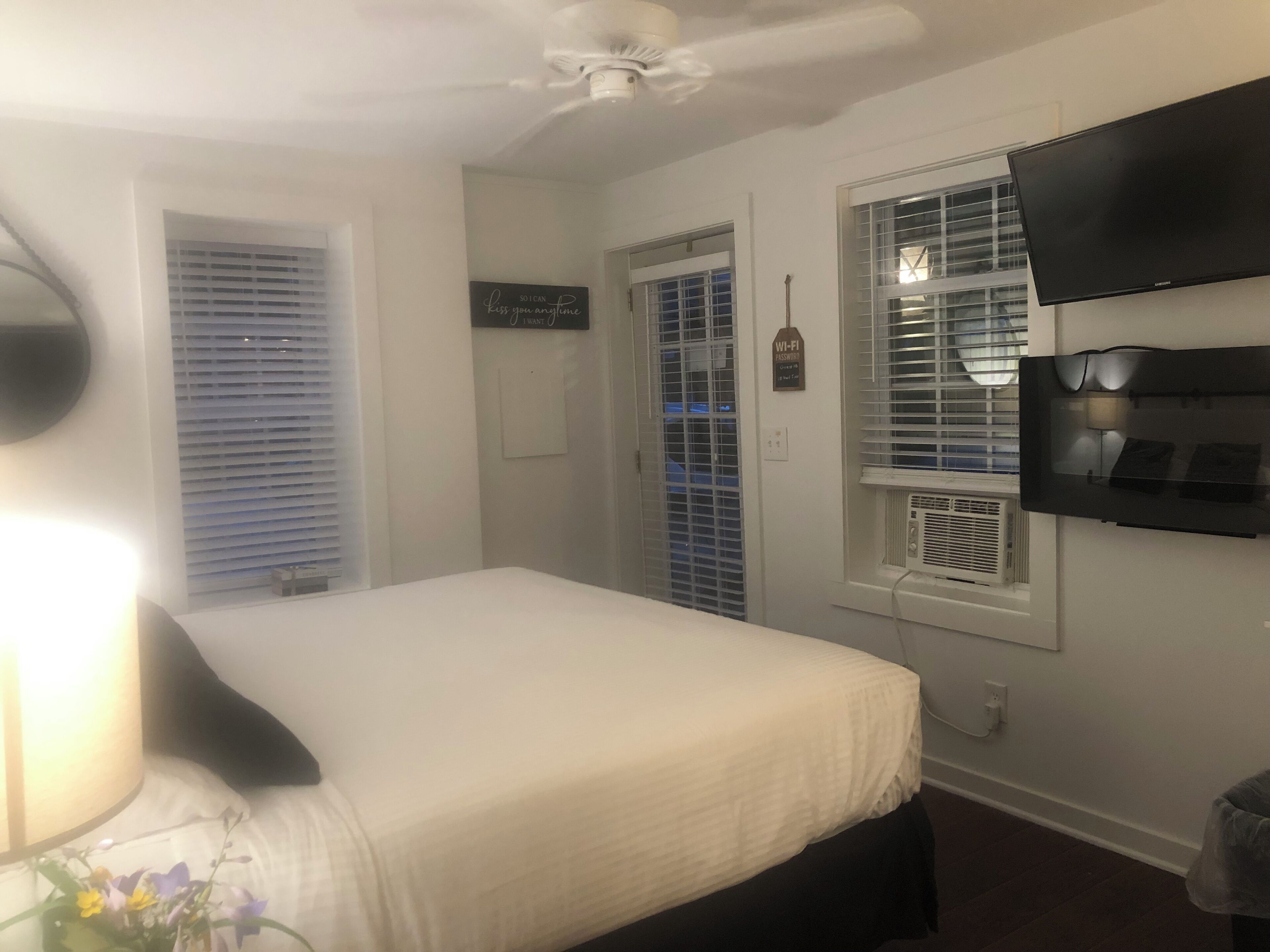 A bedroom with a bed, a ceiling fan, a TV mounted on the wall, and a window with blinds.
