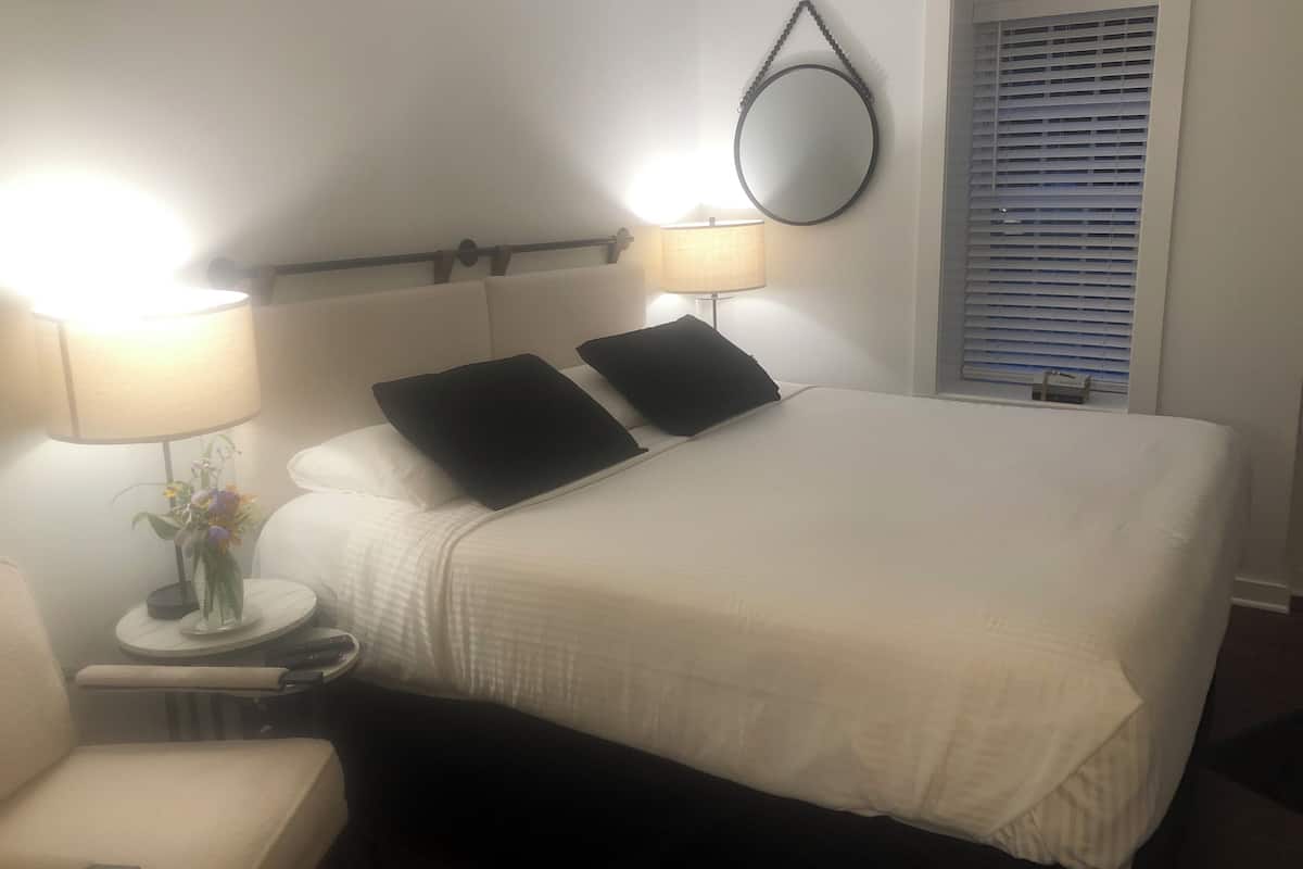 Una cama bien hecha con ropa de cama blanca y dos almohadas negras, una mesita de noche con una lámpara y un florero, y una ventana con persianas.