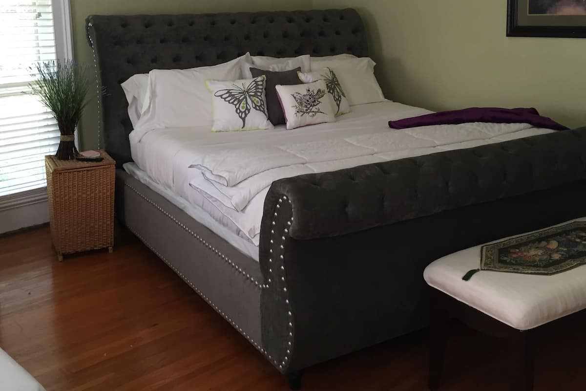 Un dormitorio con cabecera capitonada, cama grande con ropa de cama blanca, mesita de noche con planta y tres cuadros enmarcados en la pared.