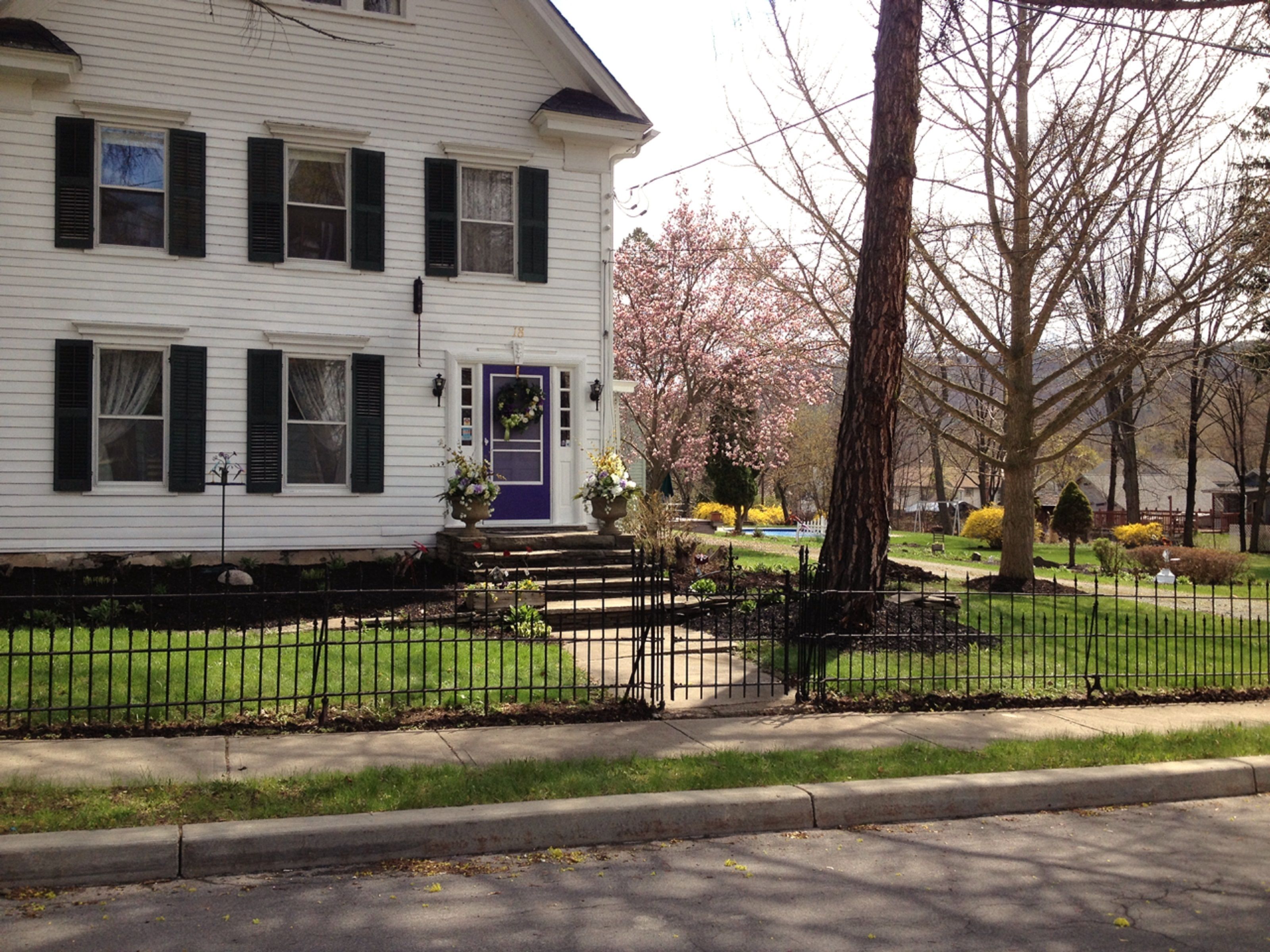 A white two-story house with black shutters, a purple front door, and a front yard with a small fence.