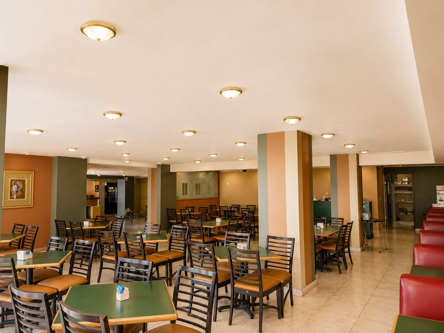 Uma sala de jantar de hotel com várias mesas e cadeiras, uma cozinha ao fundo e uma escadaria que leva a outro andar.