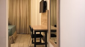 A compact hotel room with a wooden dining table, a TV mounted on the wall, a bed with a green cover, and a large mirror.