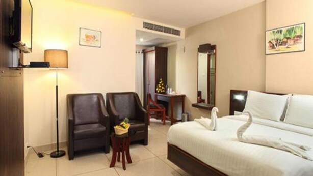 A hotel room with a bed, two armchairs, a small table, a lamp, a ceiling fan, and a TV. - Beith Hotel (Kanayannur)