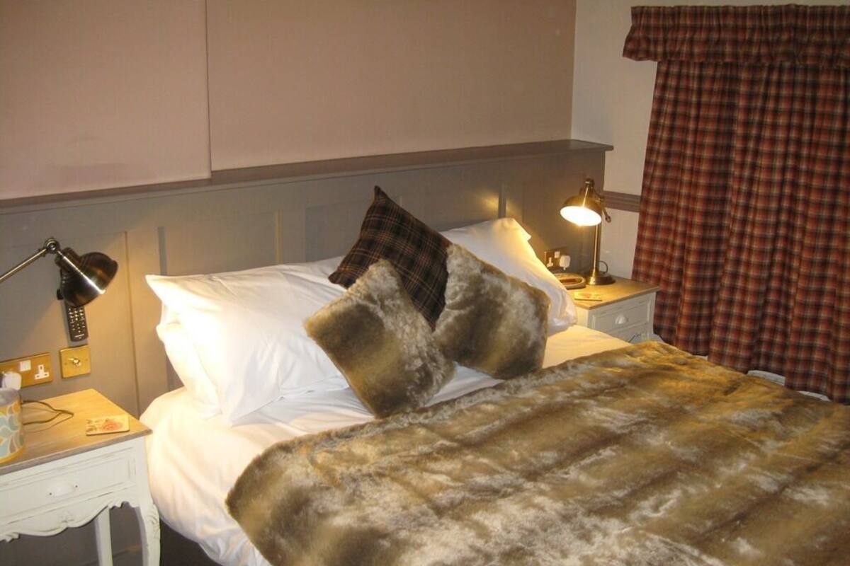 A hotel room with a bed, two bedside lamps, a nightstand, a blanket, and a plaid curtain.