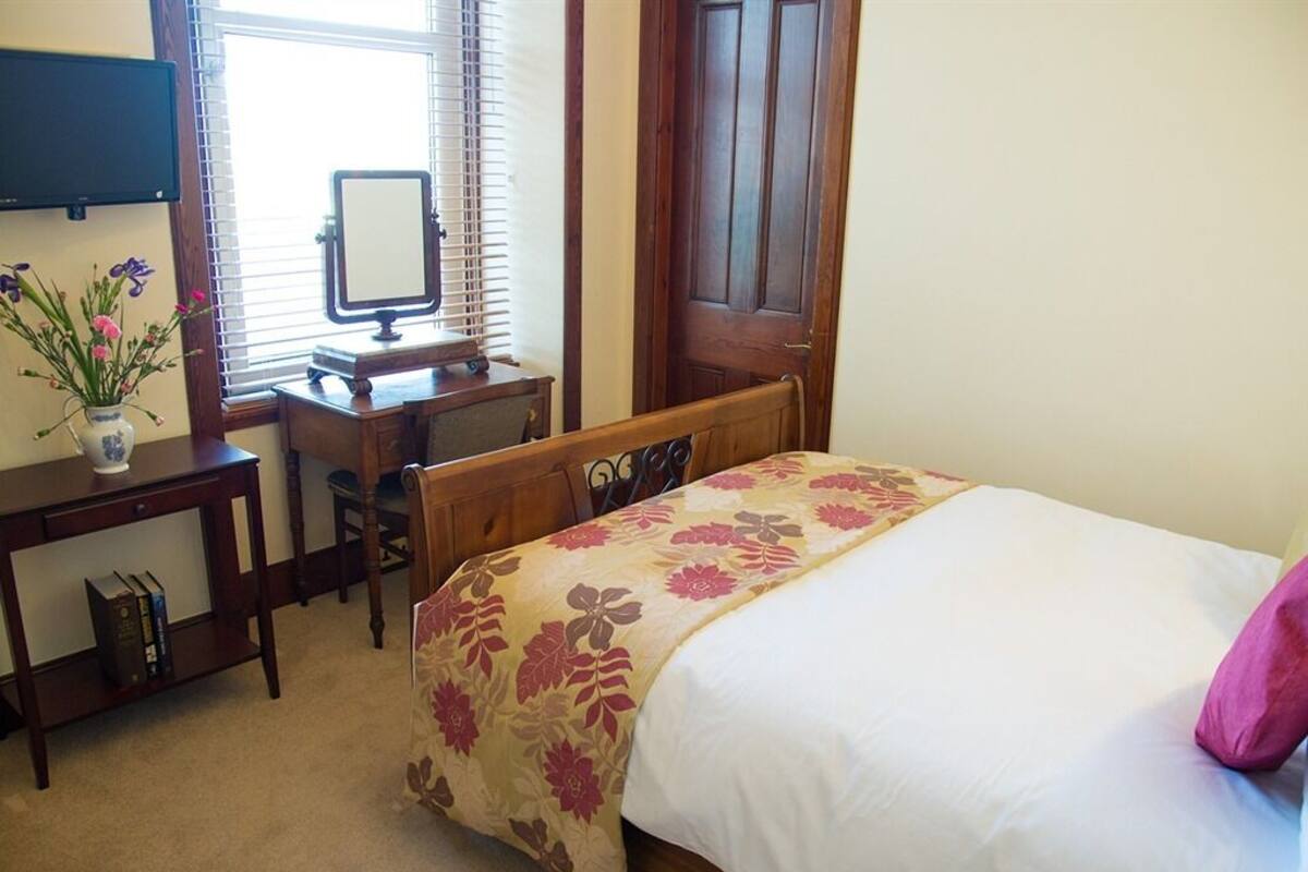 Quarto com cama, escrivaninha, cadeira, espelho e janela com persianas.