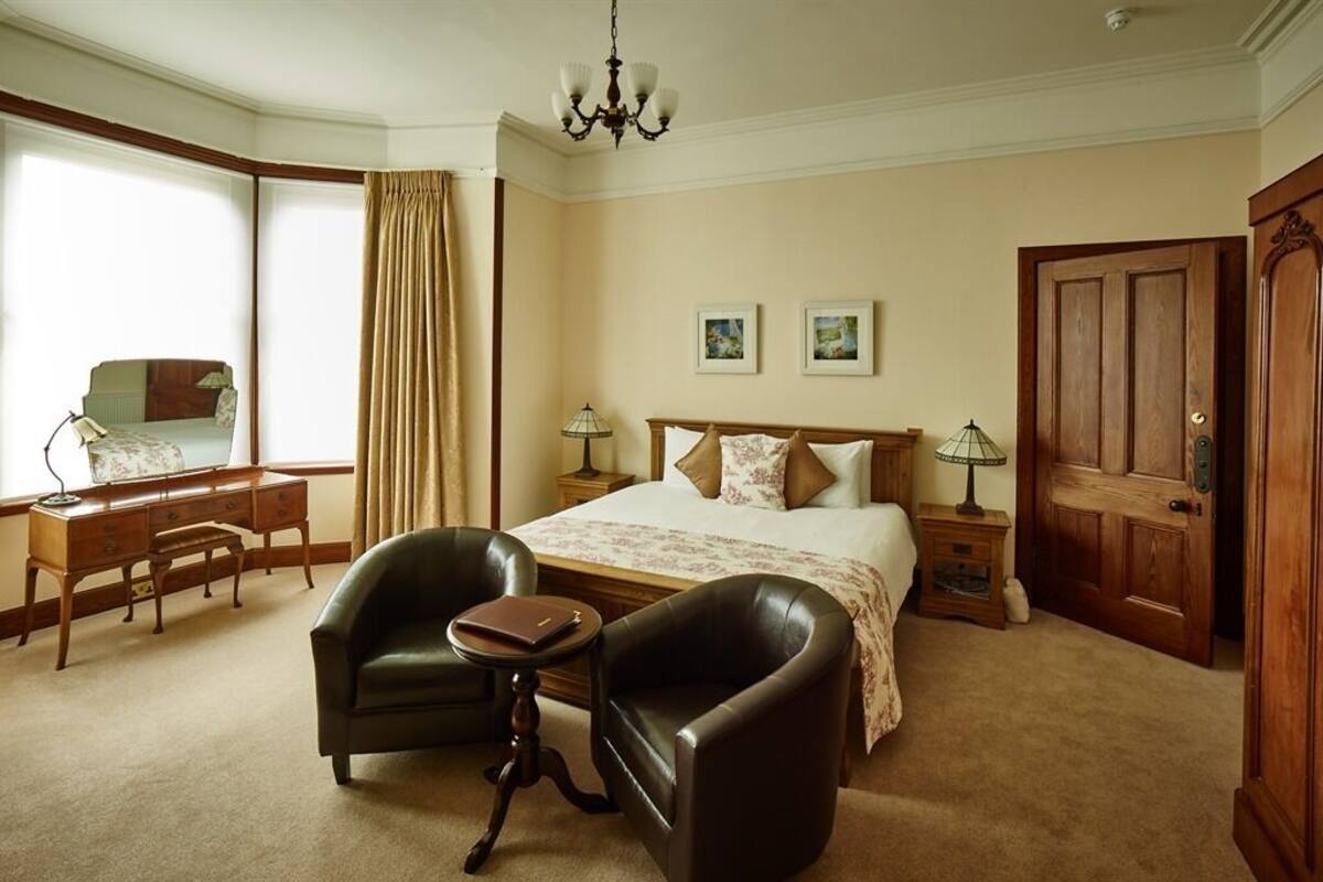Quarto de hotel com cama grande, duas luminárias de cabeceira, uma escrivaninha com cadeira, uma mesinha, uma cadeira e um guarda-roupa.