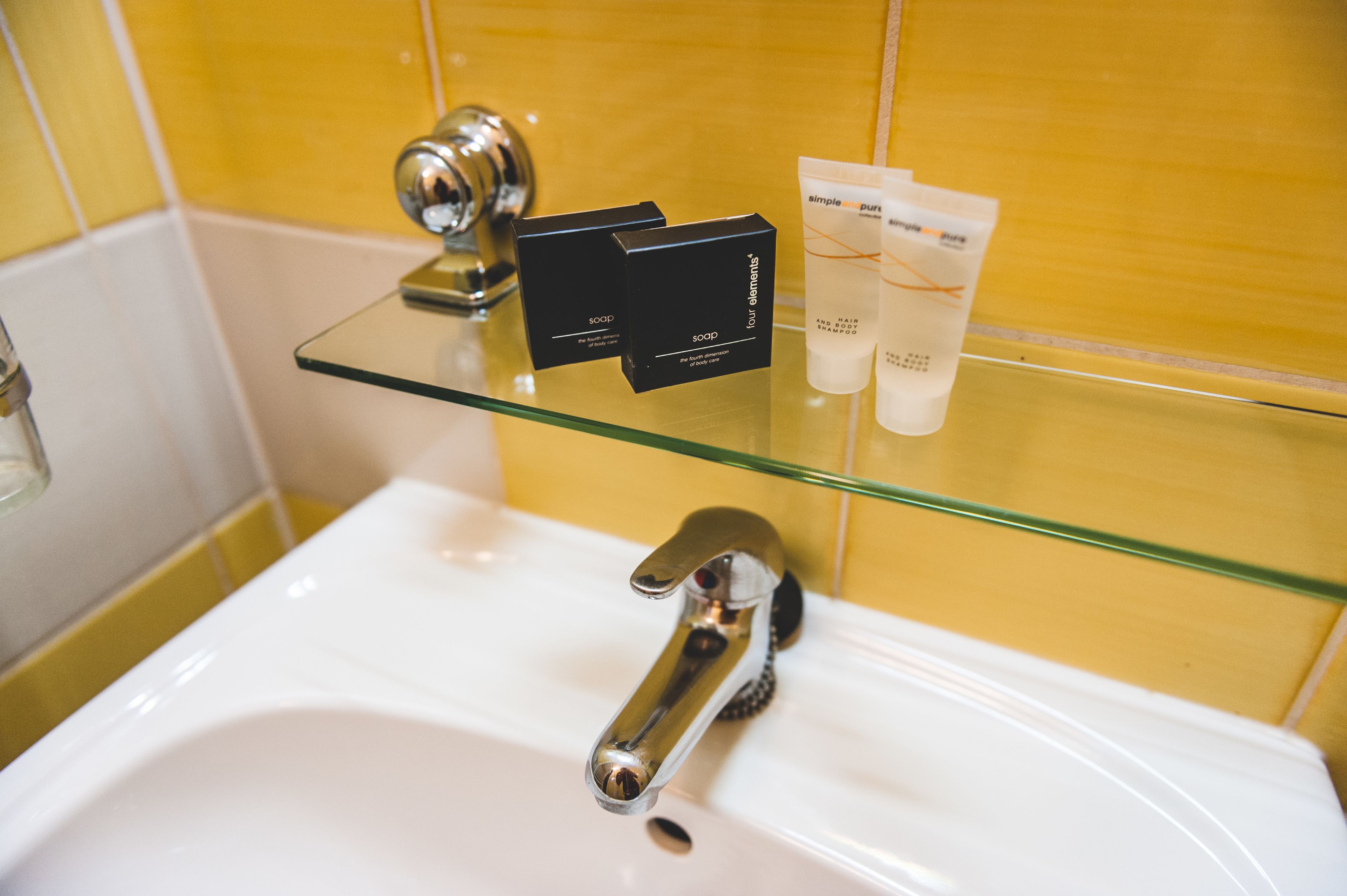 A bathroom with a glass shelf holding toiletries, a chrome faucet, and a yellow tiled wall.