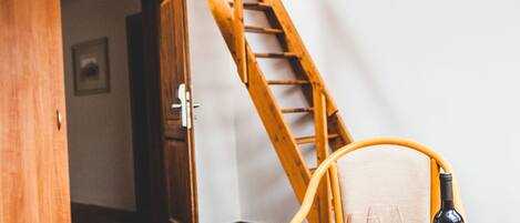 A room with a wooden staircase, a chair, a table with a bottle and glasses, and a door leading to another room.
