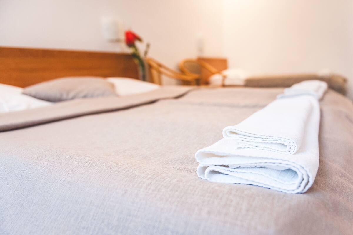 A neatly made bed with a folded white towel, a wooden headboard, and a bedside table with a vase of flowers.