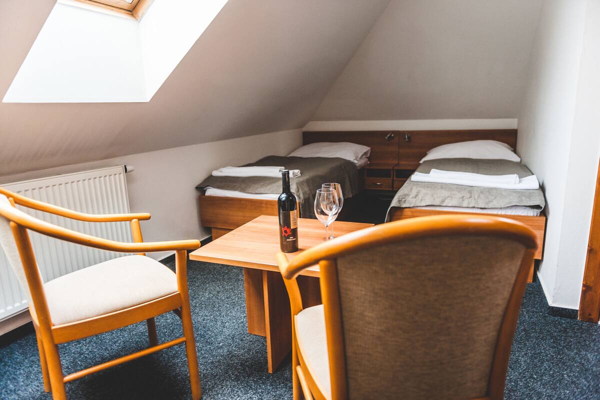 A small, cozy room with a sloped ceiling, two single beds, a wooden table with a bottle and glass, and two wooden chairs.