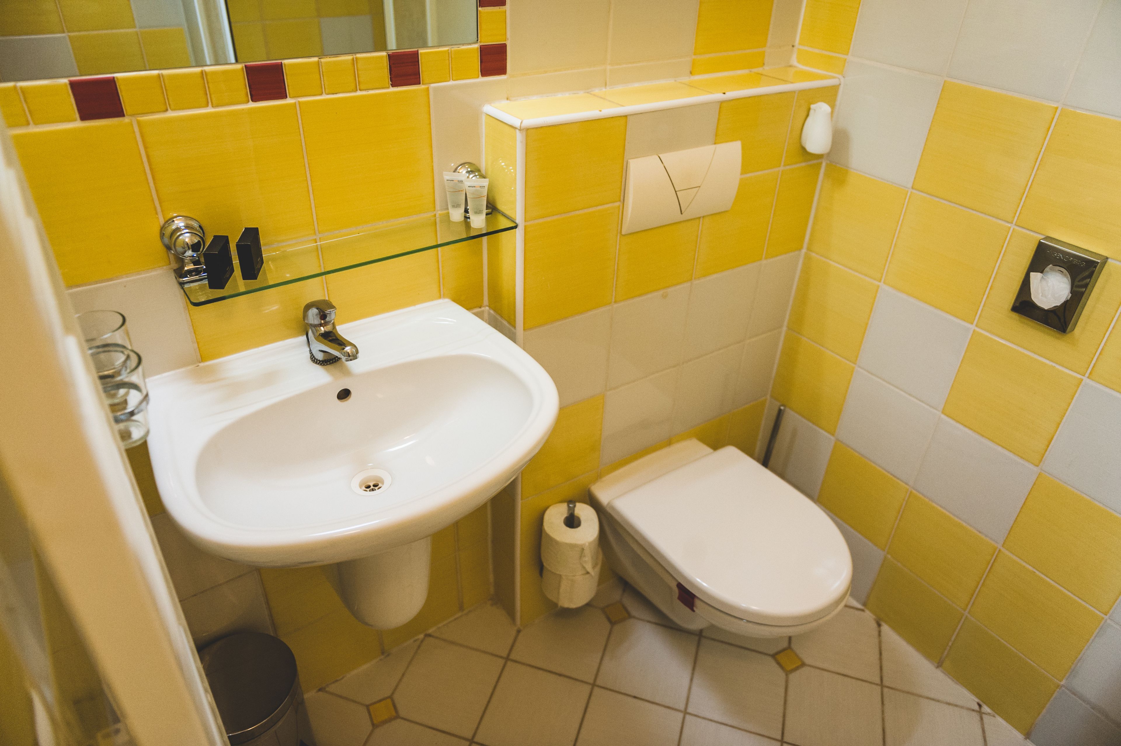 A bathroom with yellow and white tiles, a white sink, a toilet, and a glass shelf with toiletries.
