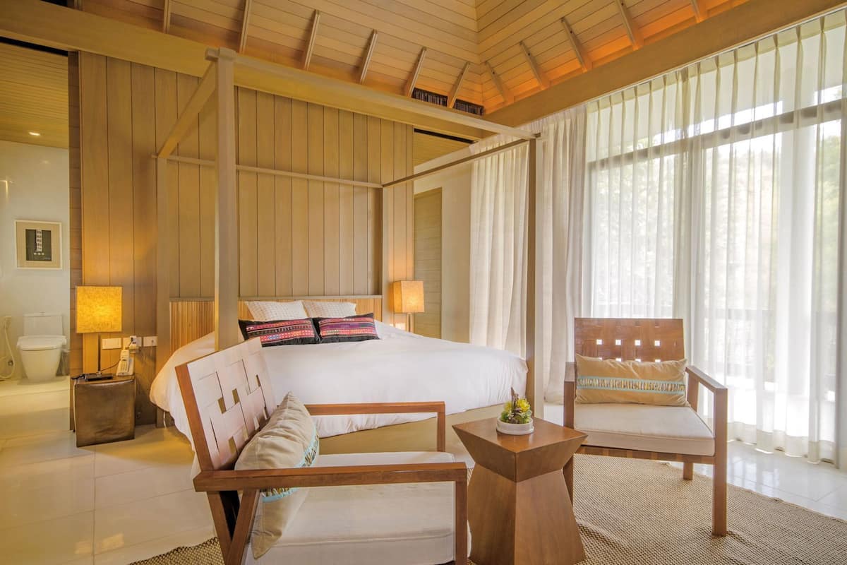 A four-poster bed with a wooden headboard, a wooden chair, a small table, and a large window with sheer curtains.