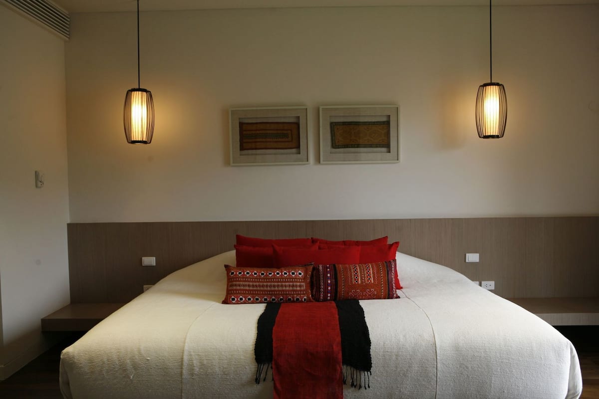 A neatly made bed with a white duvet, red and patterned pillows, and a black throw. Two wall-mounted light fixtures hang above the bed.