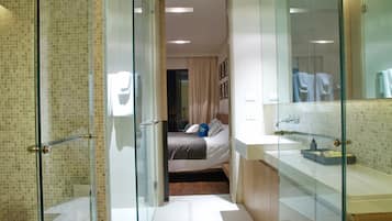 A modern hotel room with a glass shower enclosure, a bed with a gray and white duvet, and a bathroom with a large mirror and a sink.