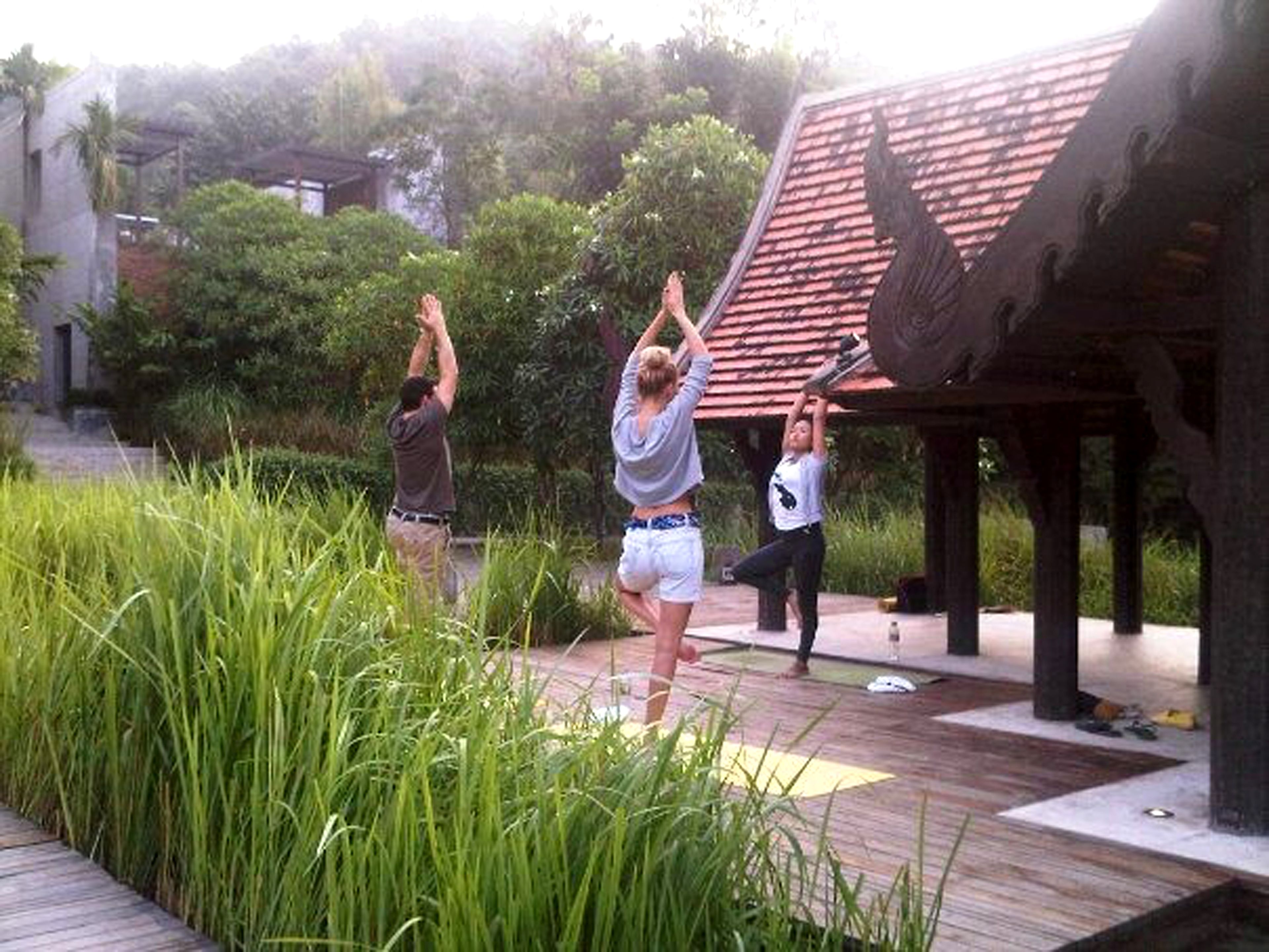 Eine Gruppe von Menschen praktiziert Yoga im Freien auf einer Holzterrasse, im Hintergrund eine traditionelle Dachkonstruktion.