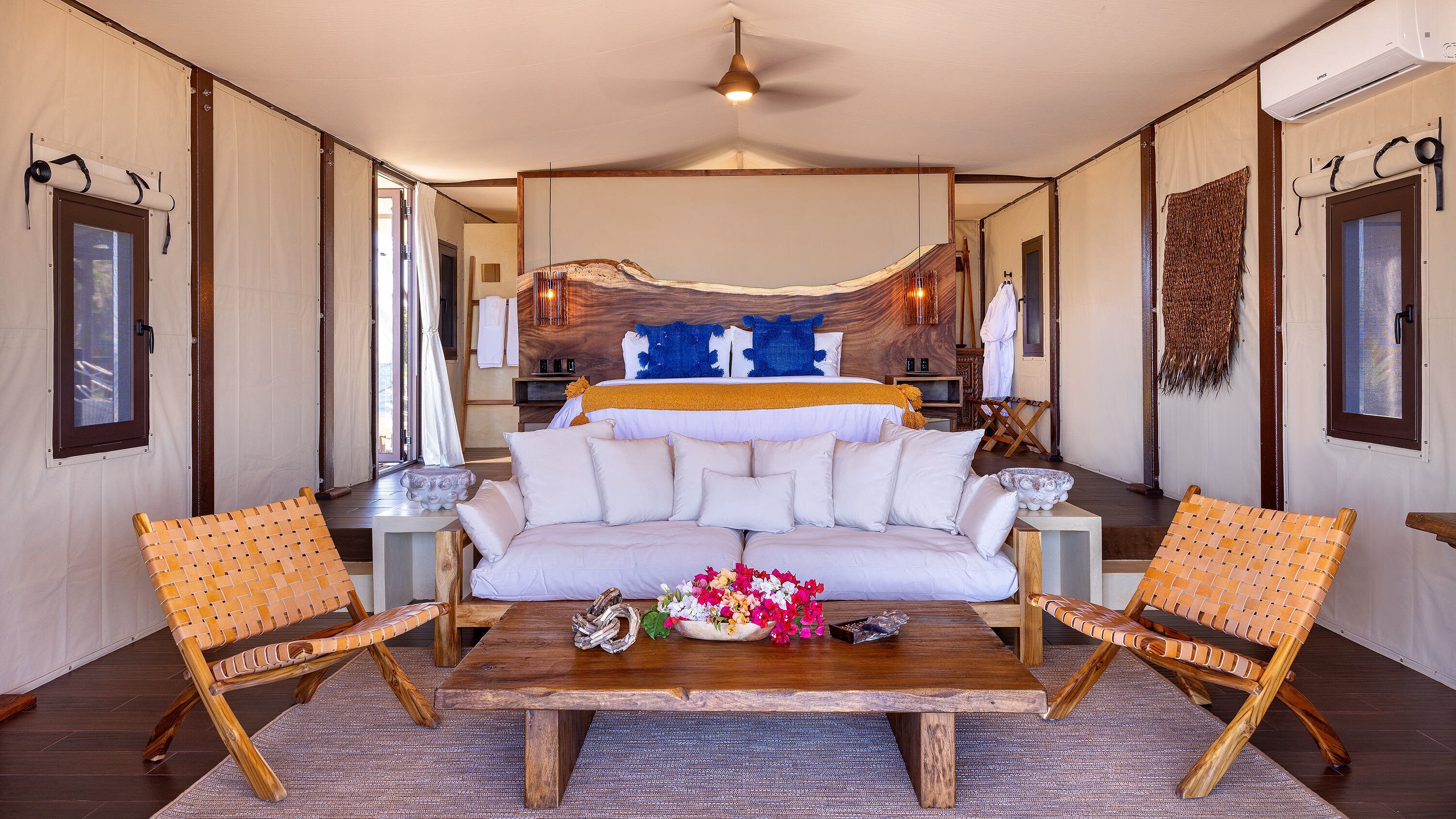 Una habitación amplia con una cama grande, dos sillas de mimbre, una mesa de centro de madera y vistas a una cabaña de madera a través de un ventanal.