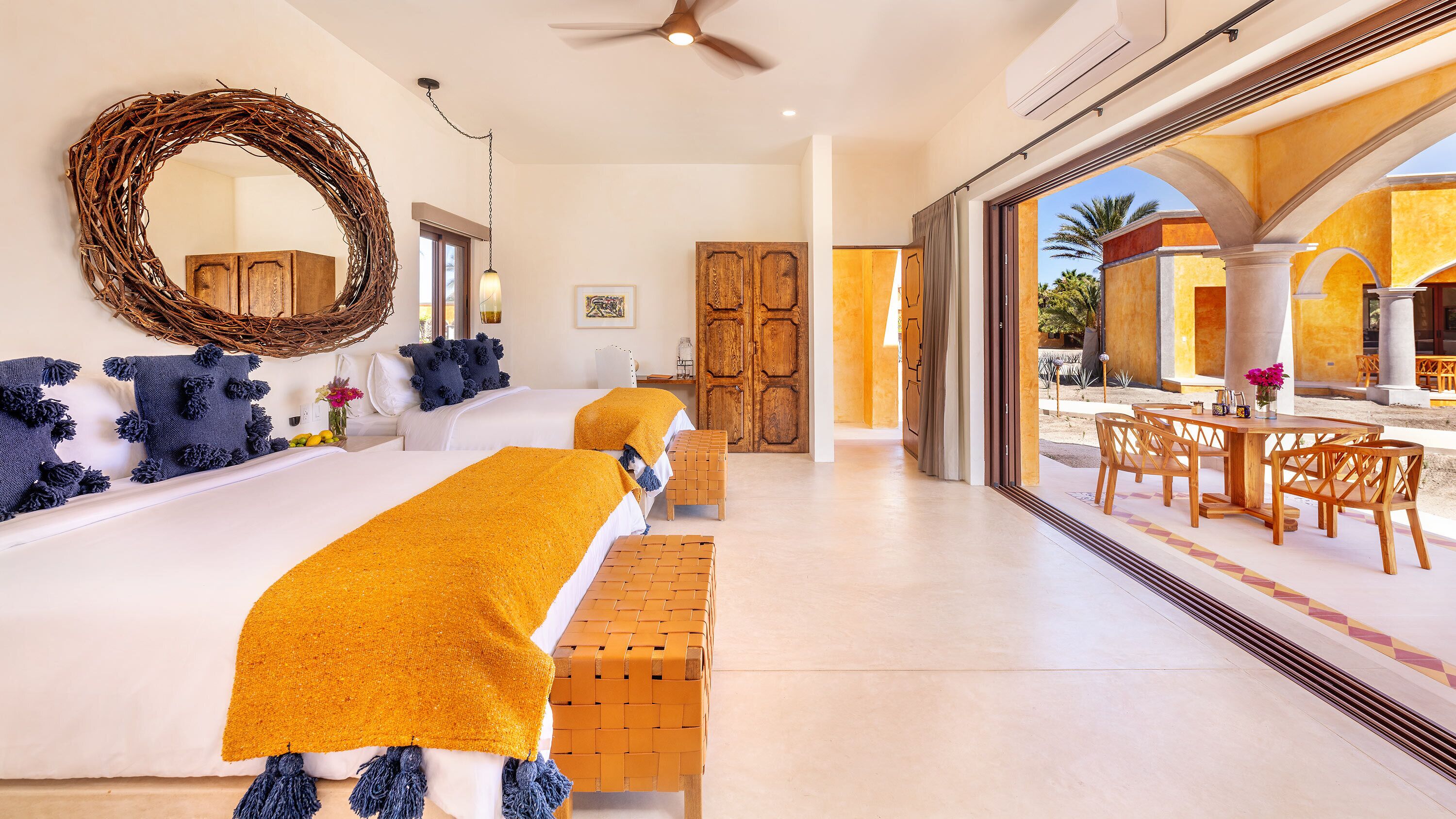 A hotel room with a large bed, a wooden door, a dining area with wooden furniture, and a view of the outdoors.