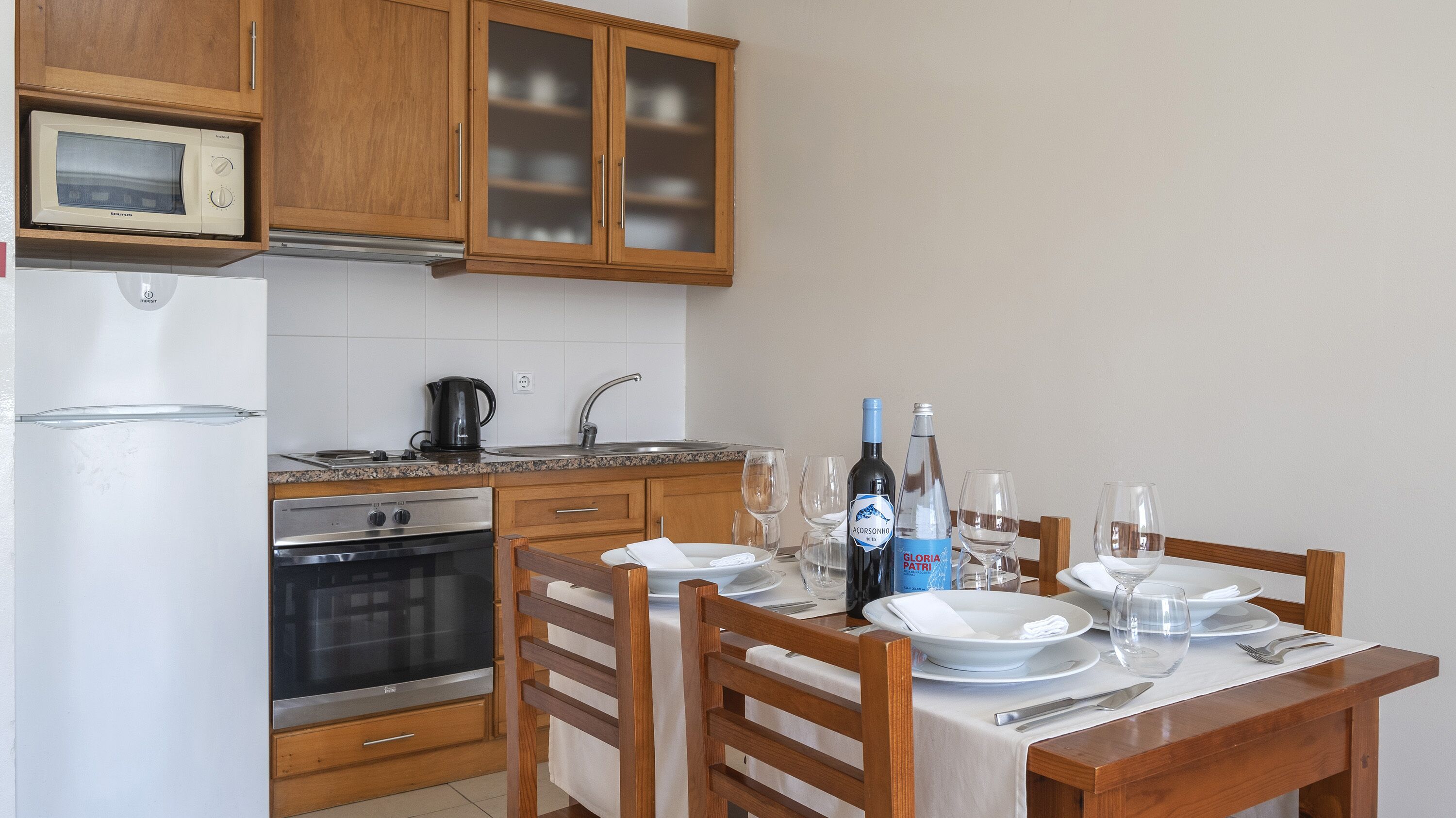 A kitchen with wooden cabinets, a microwave, a stove, and a dining table set for two with wine glasses and plates.