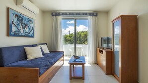 A room with a blue sofa, a small wooden table, a TV on a wooden cabinet, and a large window with a view of trees and a clear sky.