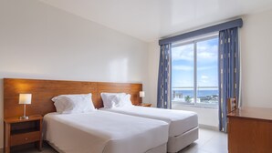 A hotel room with two beds, a wooden headboard, a nightstand with a lamp, a view of the sea through large windows, and a wooden desk.