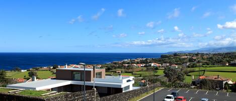 A modern building with a stone wall, a parking lot with several cars, and a view of the ocean and distant houses.