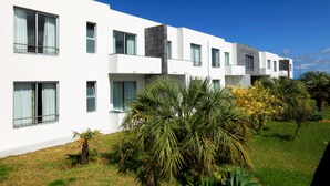 A modern hotel with a white facade, multiple balconies, and a well-maintained garden with palm trees and yellow flowering plants.