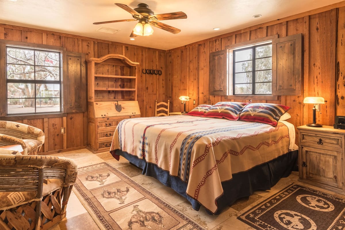 Quarto com decoração em madeira, com uma cama, duas cadeiras, uma mesa de cabeceira e um ventilador de teto.