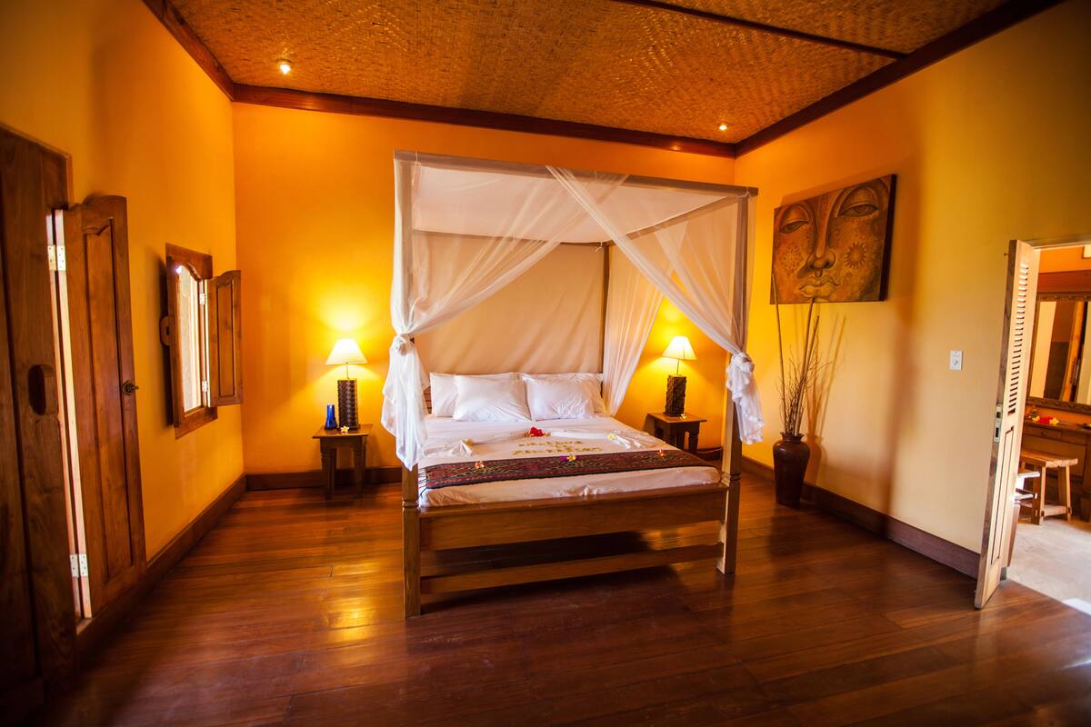 A four-poster bed with a canopy, wooden flooring, bedside lamps, and a painting on the wall.