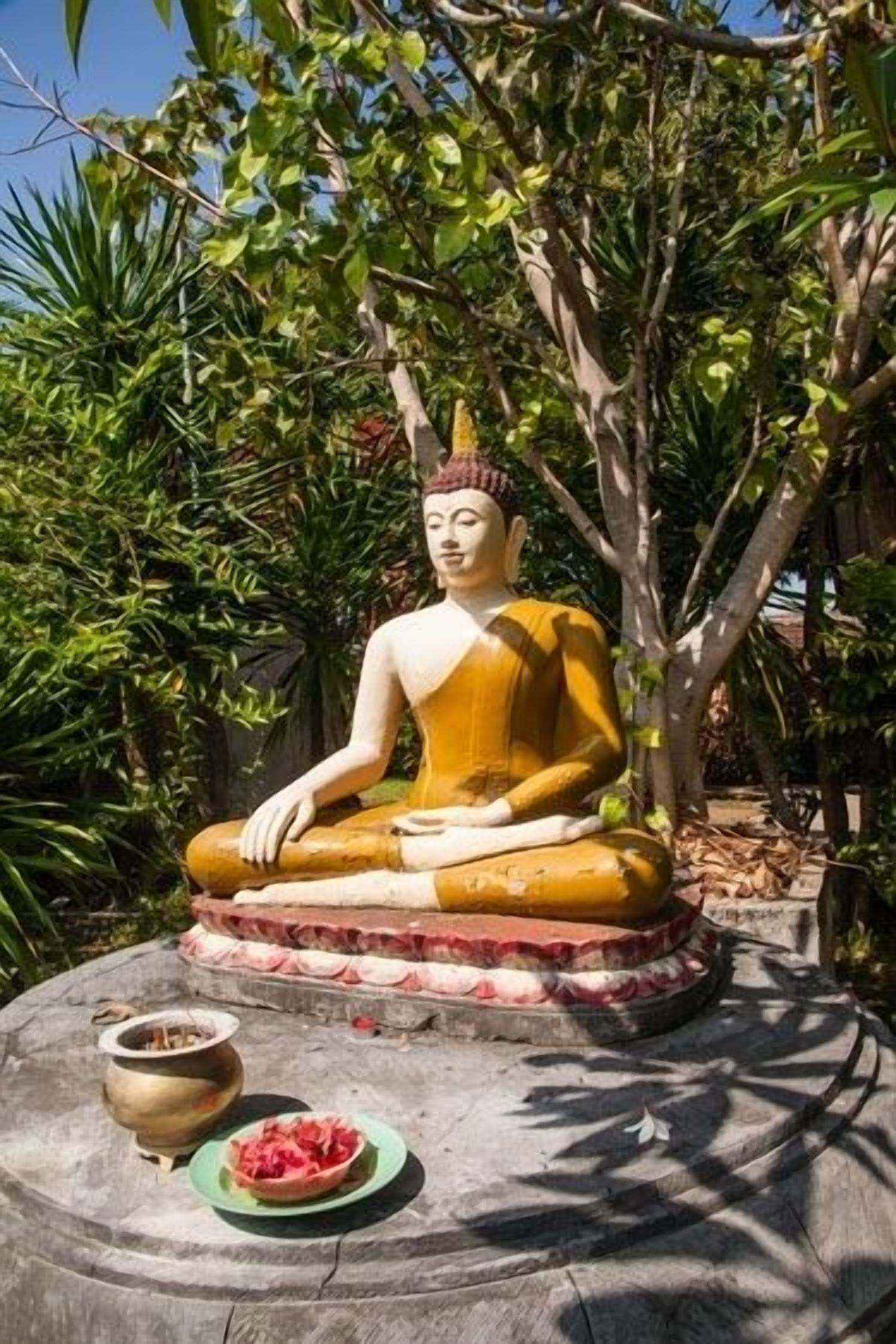 Une statue de Bouddha assis, placée sur un piédestal, avec un bol et un pot à ses côtés.