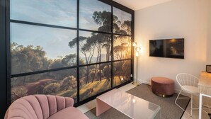 Un salon moderne doté d’une grande fenêtre, d’un téléviseur à écran plat, d’une table basse en verre, d’un canapé rose, d’un fauteuil blanc et d’un pouf rond.
