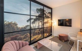 Uma sala de estar moderna com uma janela ampla, uma televisão de tela plana, uma mesa de centro de vidro, um sofá rosa, uma cadeira branca e um banquinho redondo.