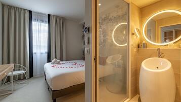 Habitación de hotel con cama, escritorio, silla, ducha y baño con un espejo grande y lavamanos.