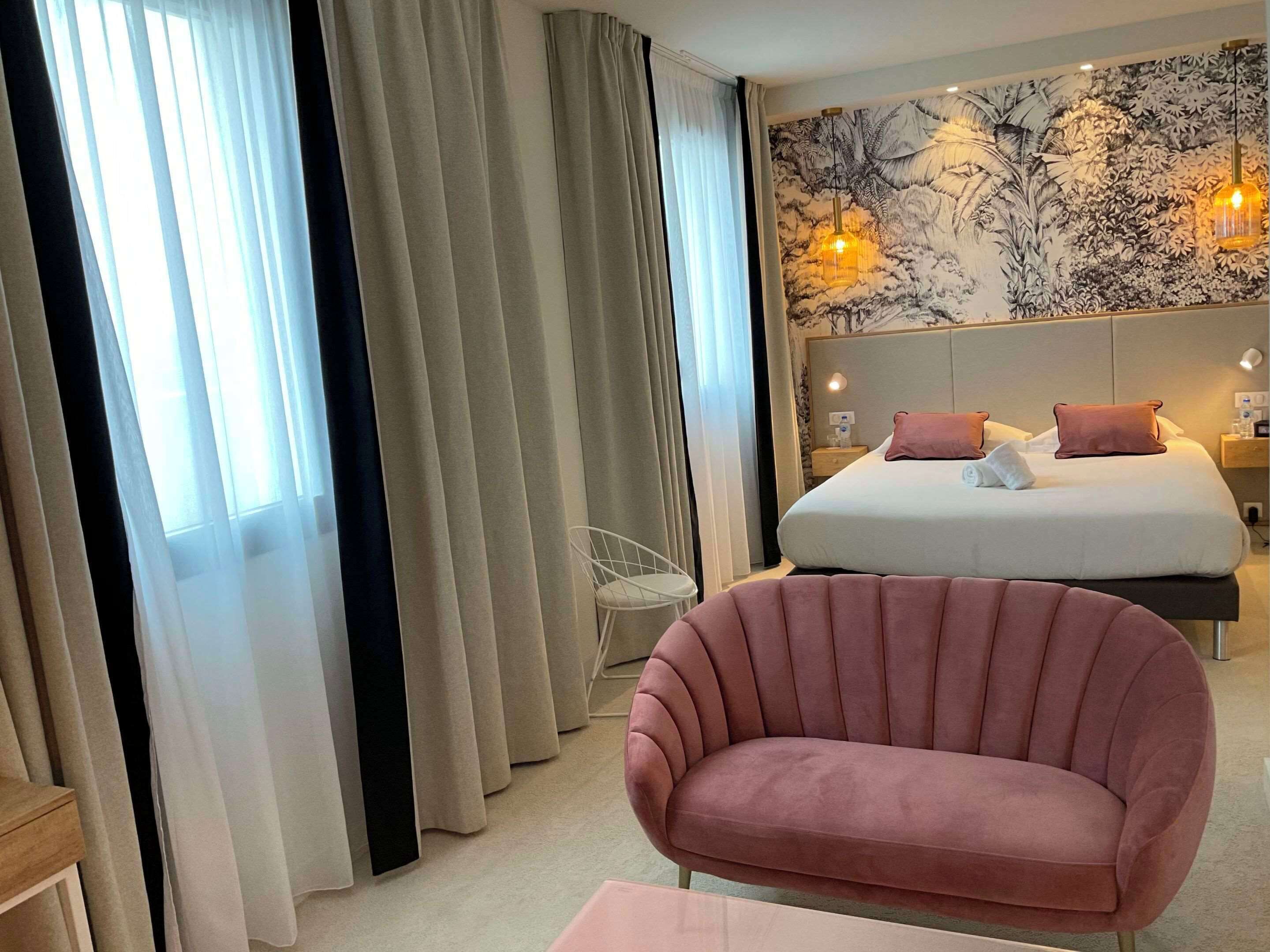 A modern hotel room with a large bed, a sitting area with a pink chair, and a wall with a floral mural.