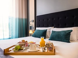 Cama bem arrumada com bandeja de itens de café da manhã, incluindo frutas, suco e iogurte.