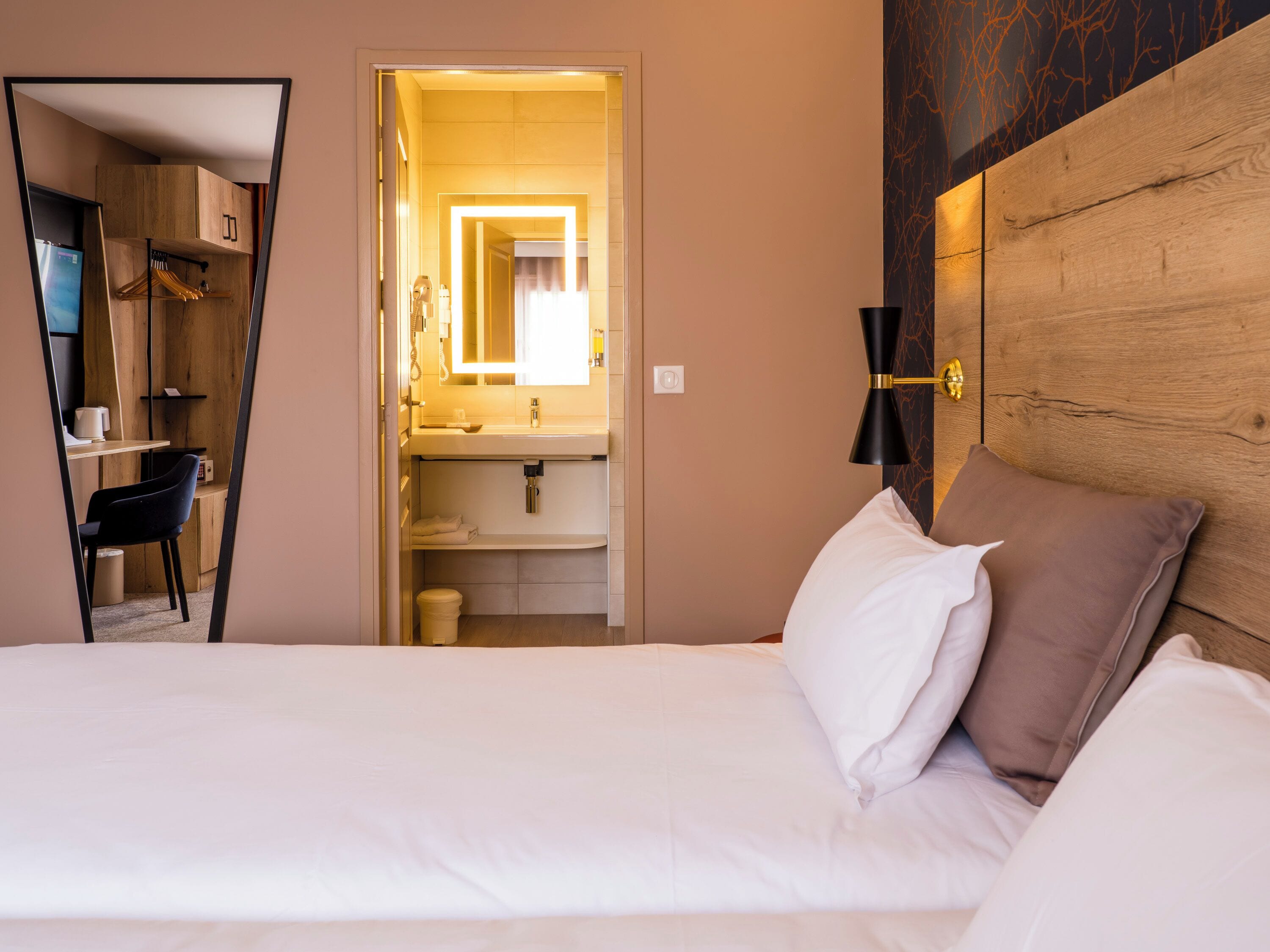 A hotel room with a bed, a desk with a chair, a mirror, and a wooden headboard.