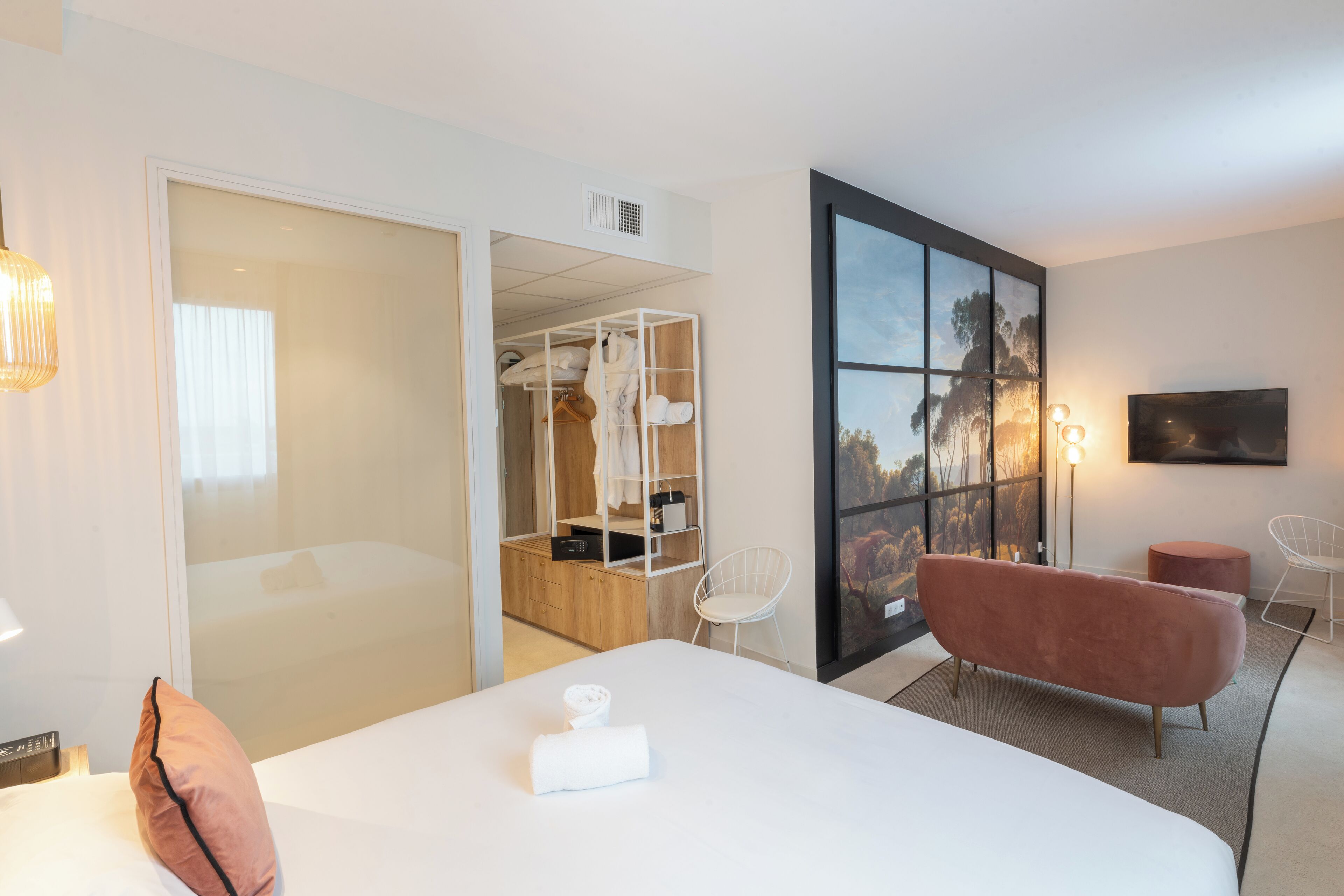 A modern hotel room with a large bed, a walk-in closet, a seating area with a pink chair, and a large window with a view.