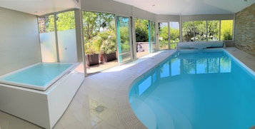 Piscina interna moderna com paredes de vidro transparente, piso ladrilhado e vista para o verde lá fora.