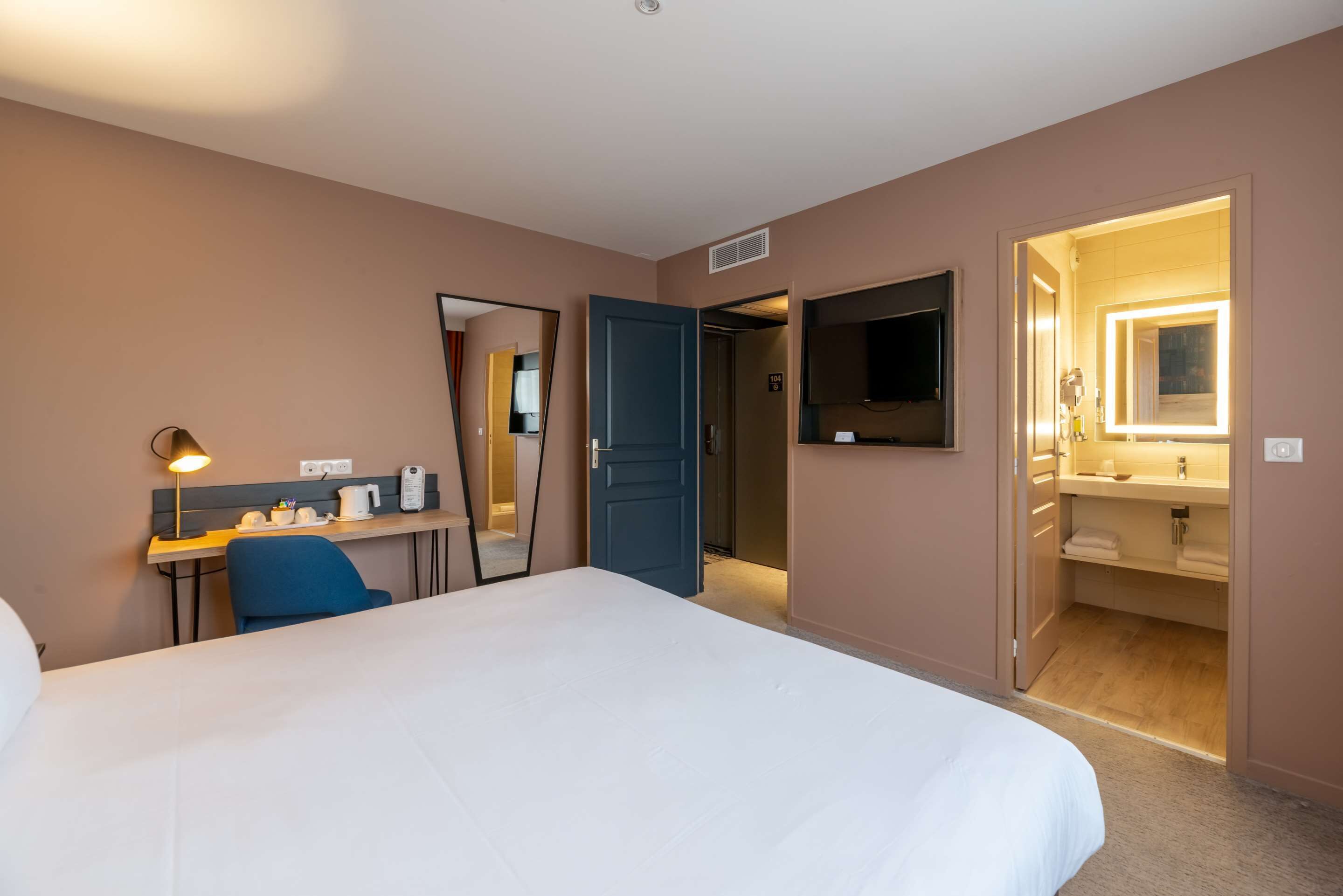 Une chambre d’hôtel avec un grand lit, un bureau avec une lampe, un miroir et une salle de bain visible à travers une porte ouverte.