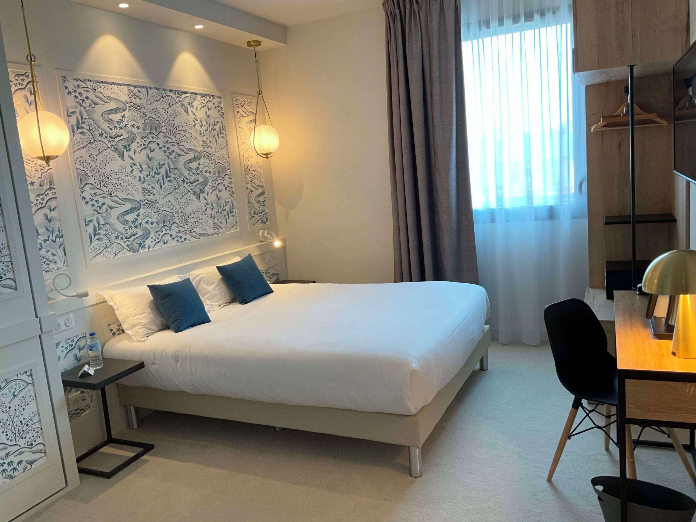 Une chambre d’hôtel moderne avec un lit bien fait, un bureau avec une chaise et une grande fenêtre avec des rideaux.