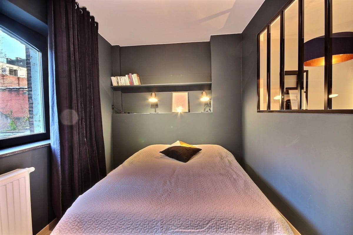 Een kamer met een bed, een raam met gordijnen, een plank met boeken en wandlampen.