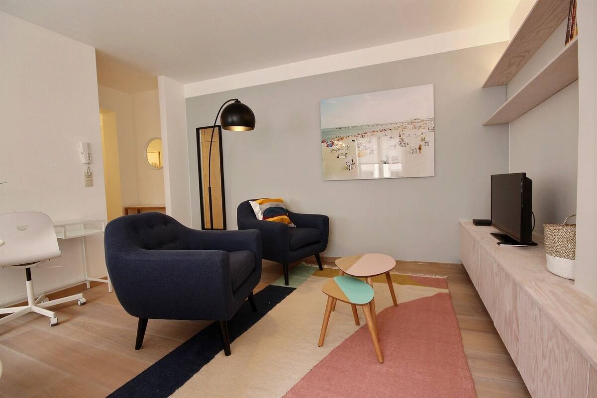Een moderne woonkamer met een bank, fauteuil, kleine tafels, een televisie en een schilderij aan de muur.