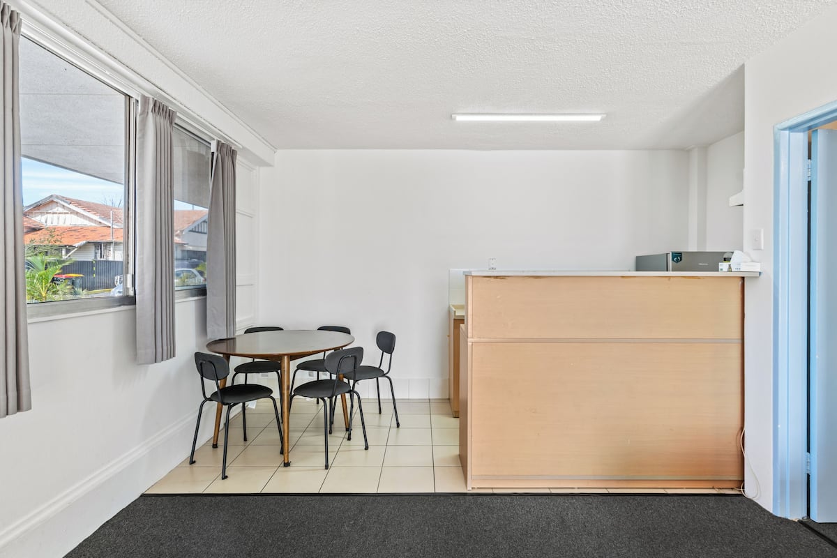 A small, clean room with a round table, four chairs, a wooden desk, and a window with curtains.