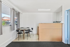 A small, clean room with a round table, four chairs, a wooden desk, and a window with curtains.
