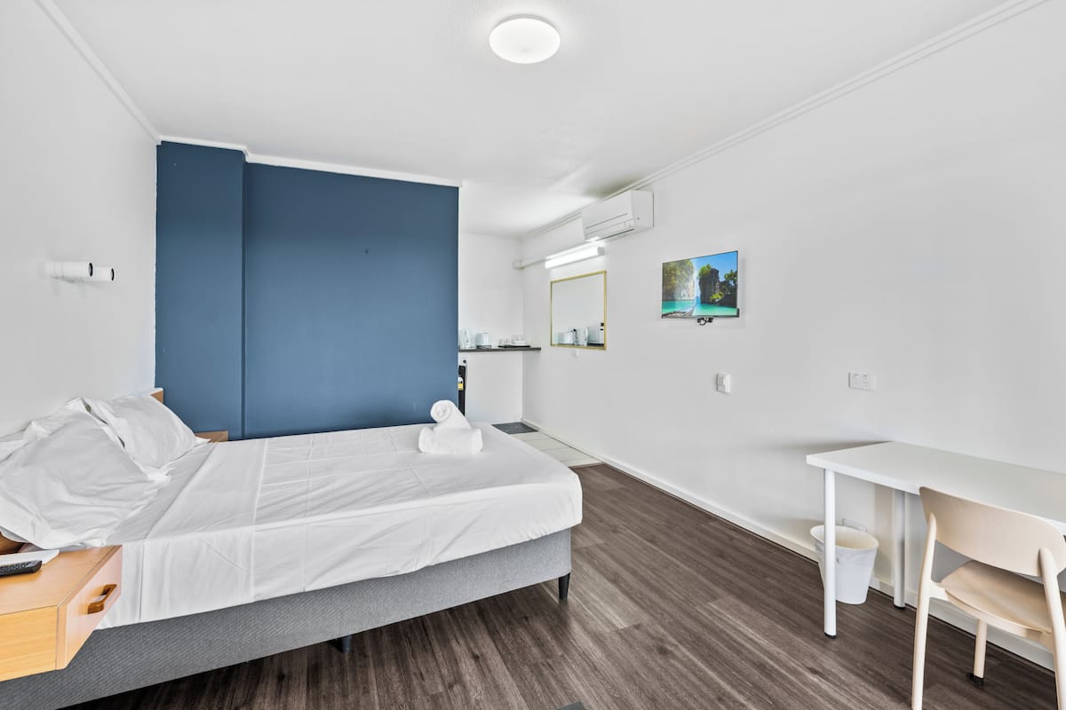 A modern hotel room with a bed, a desk, a chair, a small table, a trash bin, a mirror, and a picture on the wall.