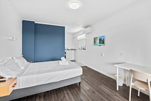 A modern hotel room with a bed, a desk, a chair, a small table, a trash bin, a mirror, and a picture on the wall.