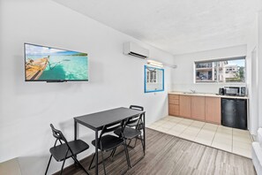 A compact room with a dining table, chairs, a TV mounted on the wall, a small kitchen area, and a window with a view of buildings.