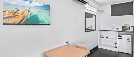 A room with a wooden table, a chair, a TV mounted on the wall, a small kitchenette, and a window.