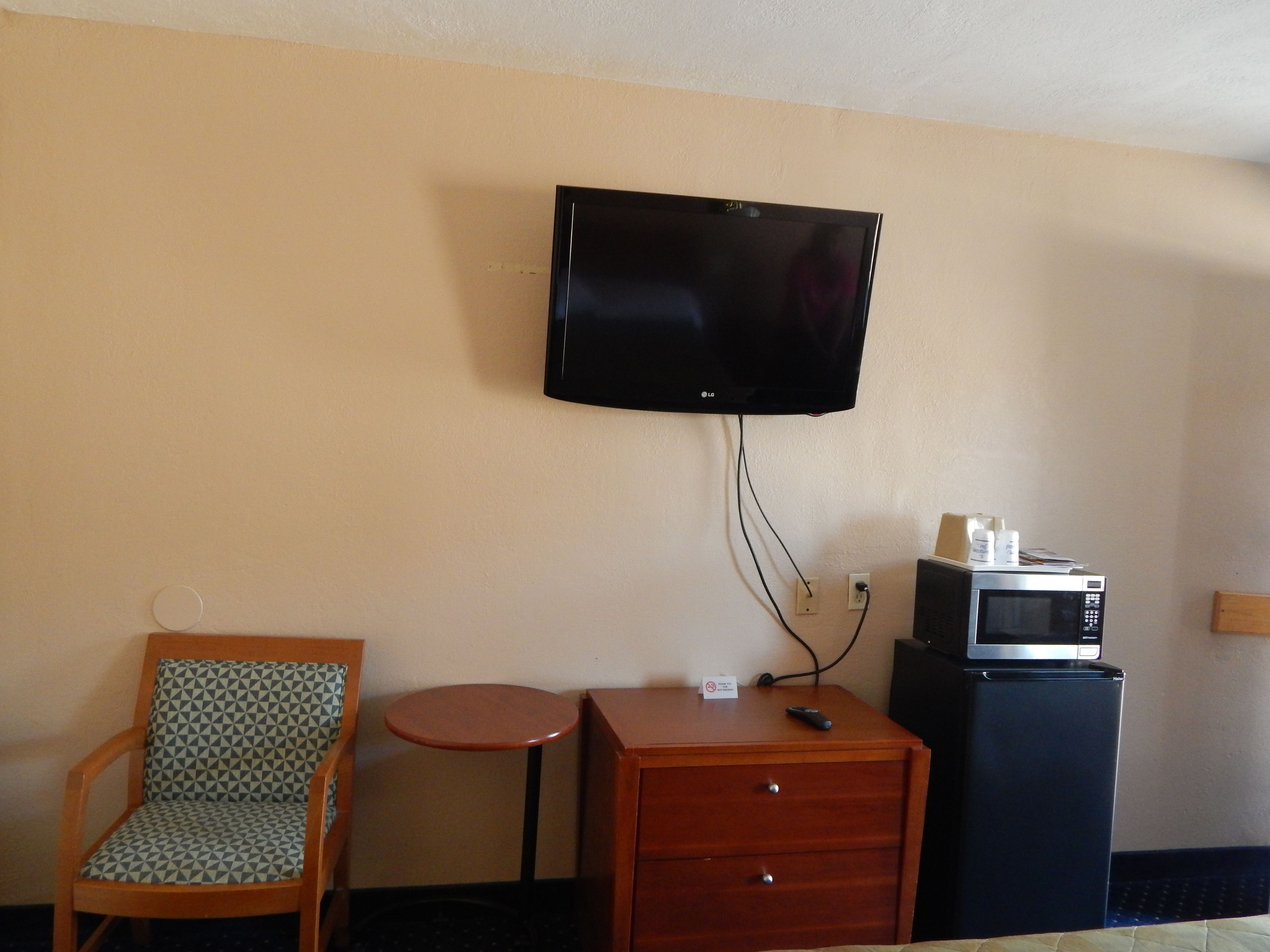 A hotel room with a television mounted on the wall, a wooden chair, a small table, a drawer unit, and a microwave.